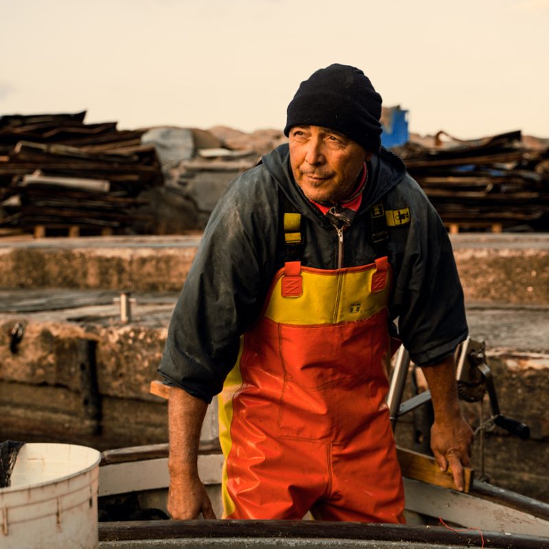 Mitilicoltore a bordo della sua imbarcazione, con abbigliamento da lavoro impermeabile, mentre opera tra attrezzature e reti per l’allevamento dei mitili.