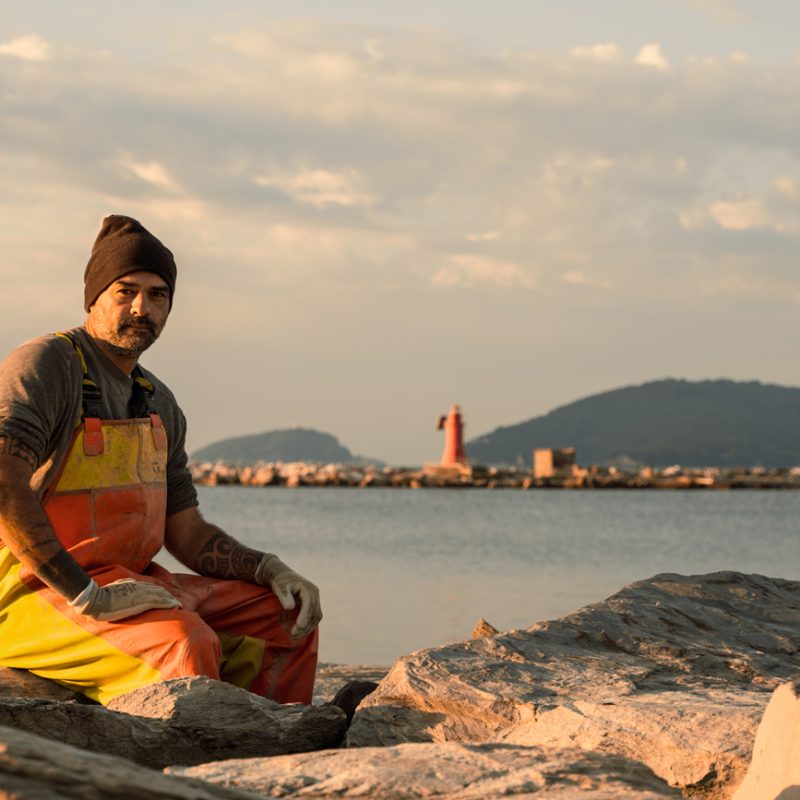Mitilicoltore seduto sugli scogli al tramonto, con indosso abiti da lavoro e il faro rosso del porto sullo sfondo.