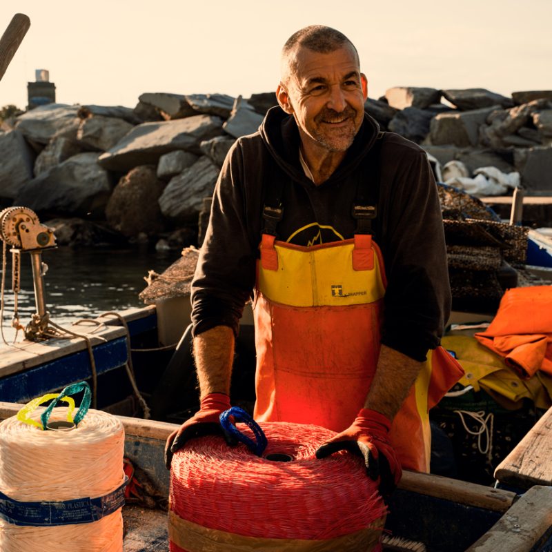 Mitilicoltore sorridente su una barca, con abbigliamento da lavoro e grandi bobine di corda utilizzate per la coltivazione dei mitili.
