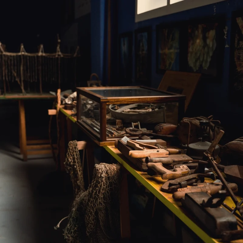 Tavolo espositivo in un museo locale con antichi strumenti da lavoro in legno e metallo, reti da pesca appese e teche contenenti reperti, illuminato da una luce calda proveniente dall’alto.