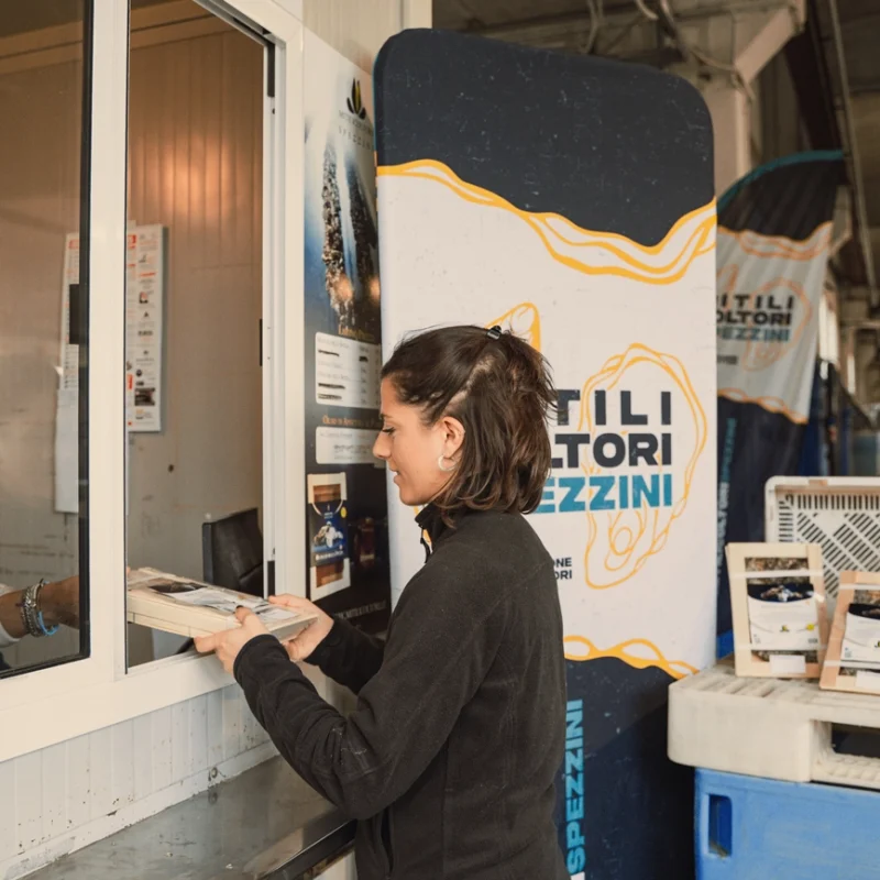 Una donna consegna confezioni di prodotti ittici attraverso una finestra di vendita a un operatore interno; accanto alla postazione sono visibili sacchi di mitili e un grande banner dei Mitilicoltori Spezzini.